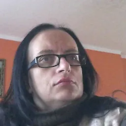 Portret kobiety w okularach na tle pomarańczowej ściany z obrazem wiatraka i lampą sufitową.