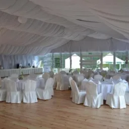 Uroczyste wnętrze namiotu bankietowego z drewnianą podłogą, białymi krzesłami z pokrowcami, okrągłymi stołami z białymi obrusami i bufetem w tle, oświetlone naturalnym światłem.