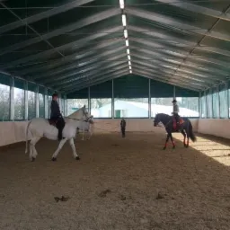 Kryta ujeżdżalnia koni z konstrukcją stalową, widoczne konie z jeźdźcami w trakcie treningu, oświetlenie jarzeniowe pod dachem.