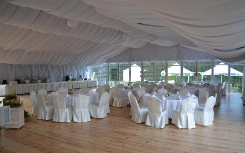 Uroczyste wnętrze namiotu bankietowego z drewnianą podłogą, białymi krzesłami z pokrowcami, okrągłymi stołami z białymi obrusami i bufetem w tle, oświetlone naturalnym światłem.