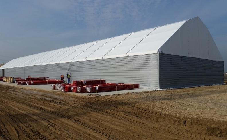 Długa hala z szarą, metalową konstrukcją ścian i białym dachem z plandeki, w trakcie montażu, z widocznymi elementami konstrukcyjnymi i pracownikami na drabinie.