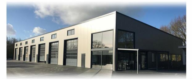 Nowoczesna hala magazynowa z szarej płyty warstwowej, widoczne bramy segmentowe i duże okna, błękitne niebo z chmurami w tle.