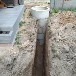 Wykop pod szambo z betonowych kręgów, widoczny z poziomu gruntu, obok fragmentu tarasu i ogrodzenia.