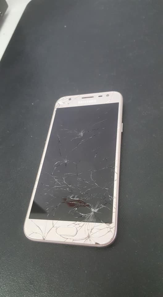 Uszkodzony smartfon Samsung z pękniętym ekranem, widoczne liczne pajęczyny pęknięć na wyświetlaczu, leżący na ciemnym, matowym tle.