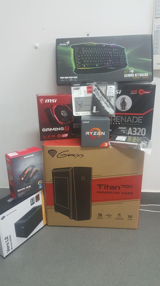 Pudełka z podzespołami komputerowymi ułożone jedno na drugim: obudowa Genesis Titan 700, procesor AMD Ryzen, karta graficzna MSI Gaming X, pamięć RAM Ballistix Sport, płyta główna Grenade A320...