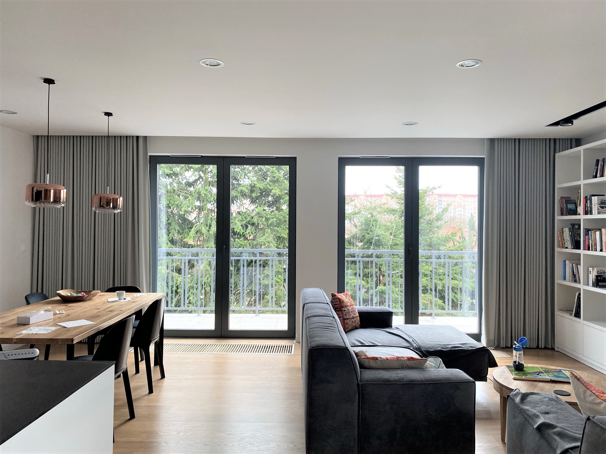 Nowoczesne wnętrze salonu z szarymi zasłonami, drewnianym stołem i czarnymi krzesłami oraz narożną sofą, widok na balkon z zielenią w tle.