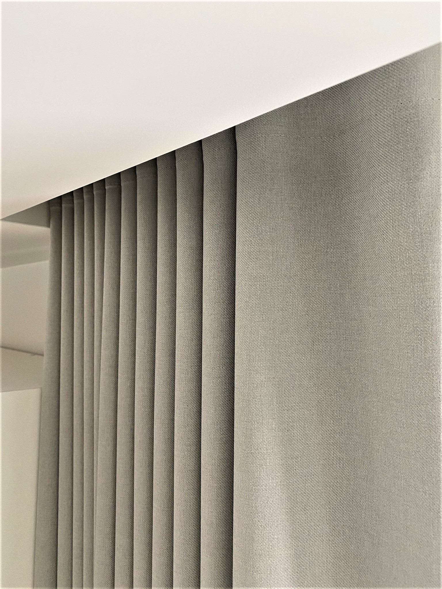 Szara, tekstylna osłona okienna z regularnymi pionowymi fałdami, zamontowana pod białym sufitem, widok z bliska.