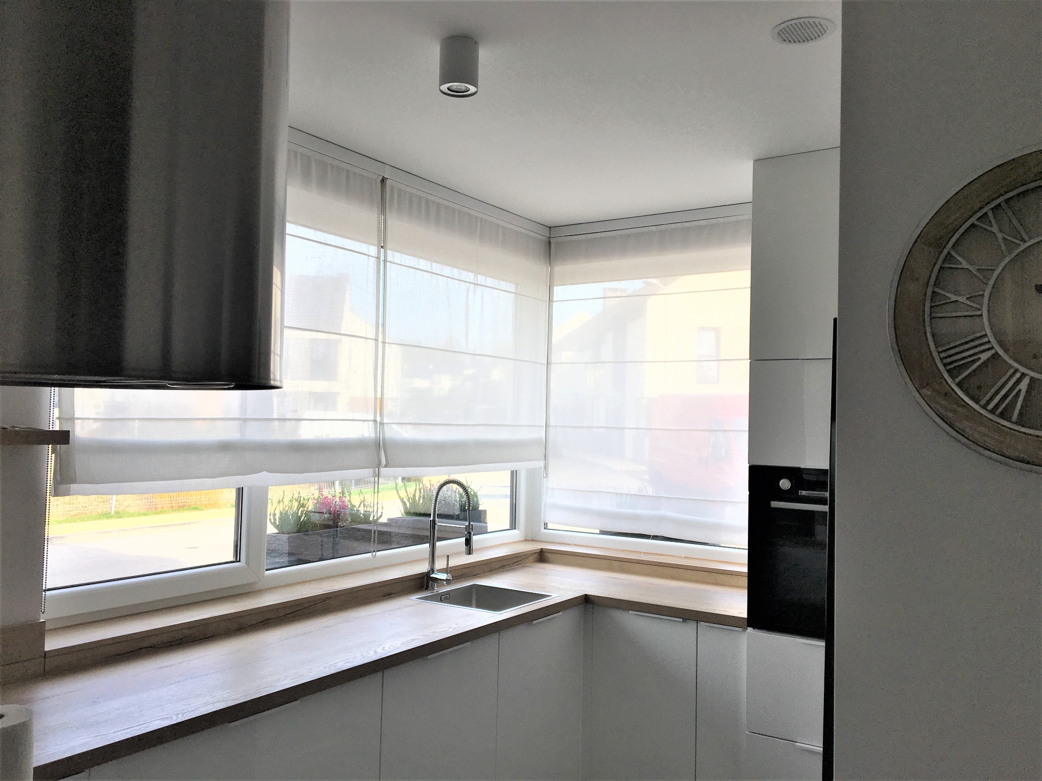 Jasna kuchnia z białymi, zwiniętymi roletami w oknach narożnych, drewnianym blatem, metalowym zlewem i minimalistycznym wystrojem, z widokiem na zieleń za oknem.