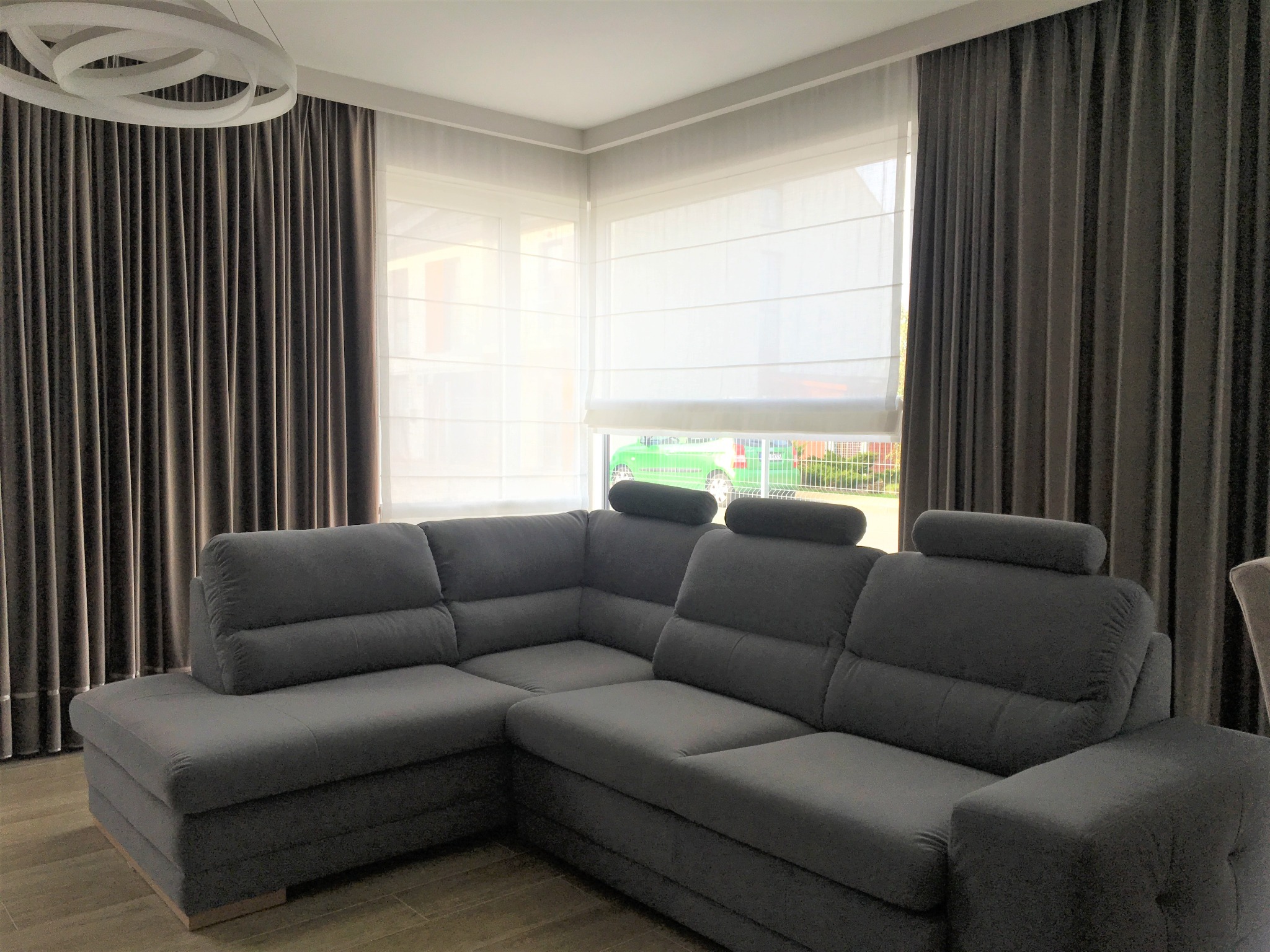 Szary narożnik z regulowanymi zagłówkami w salonie z brązowymi zasłonami i białymi roletami rzymskimi.