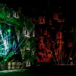 Scena koncertowa z artystką w niebieskiej sukience, intensywne zielone i czerwone oświetlenie, wizualizacje drzew z portretami w tle, zespół muzyczny w lewym rogu.