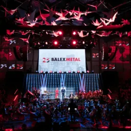 Scena z orkiestrą, podświetlona na czerwono, z dekoracjami w kształcie origami i projekcją logo 'BalexMetal' na ścianach podczas wydarzenia firmowego.