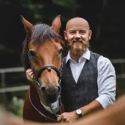 Portret uśmiechniętego mężczyzny z brodą w eleganckiej kamizelce, pozującego z brązowym koniem z uprzężą na tle zieleni.