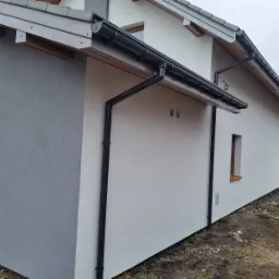 Fragment nowo otynkowanego domu jednorodzinnego z czarnymi rynnami, szarą elewacją boczną i białą elewacją frontową, widoczne otwory wentylacyjne i małe okno.