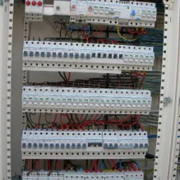 Otwarta szafa rozdzielcza z zamontowanymi bezpiecznikami i okablowaniem, widoczne przewody w kolorach czerwonym, niebieskim, żółtym i czarnym, ułożone w poziomych rzędach.