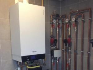 Biały piec grzewczy Viessmann zamontowany na szarej, kafelkowanej ścianie. Widoczne miedziane rury instalacji grzewczej z zaworami i manometrami. Żółty przewód gazowy doprowadzony do pieca.