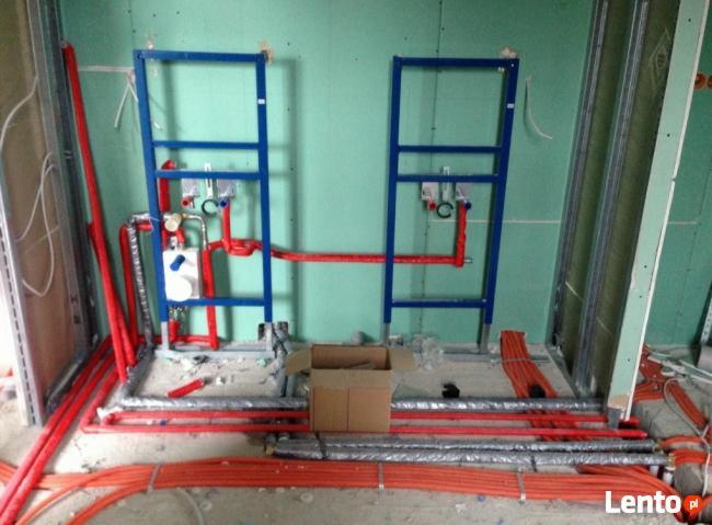 Wnętrze w trakcie instalacji wodno-kanalizacyjnej: widoczne niebieskie ramy montażowe, czerwone rury grzewcze i biały zbiornik, częściowo zakryte kartonem, na tle surowej ściany z płyt...