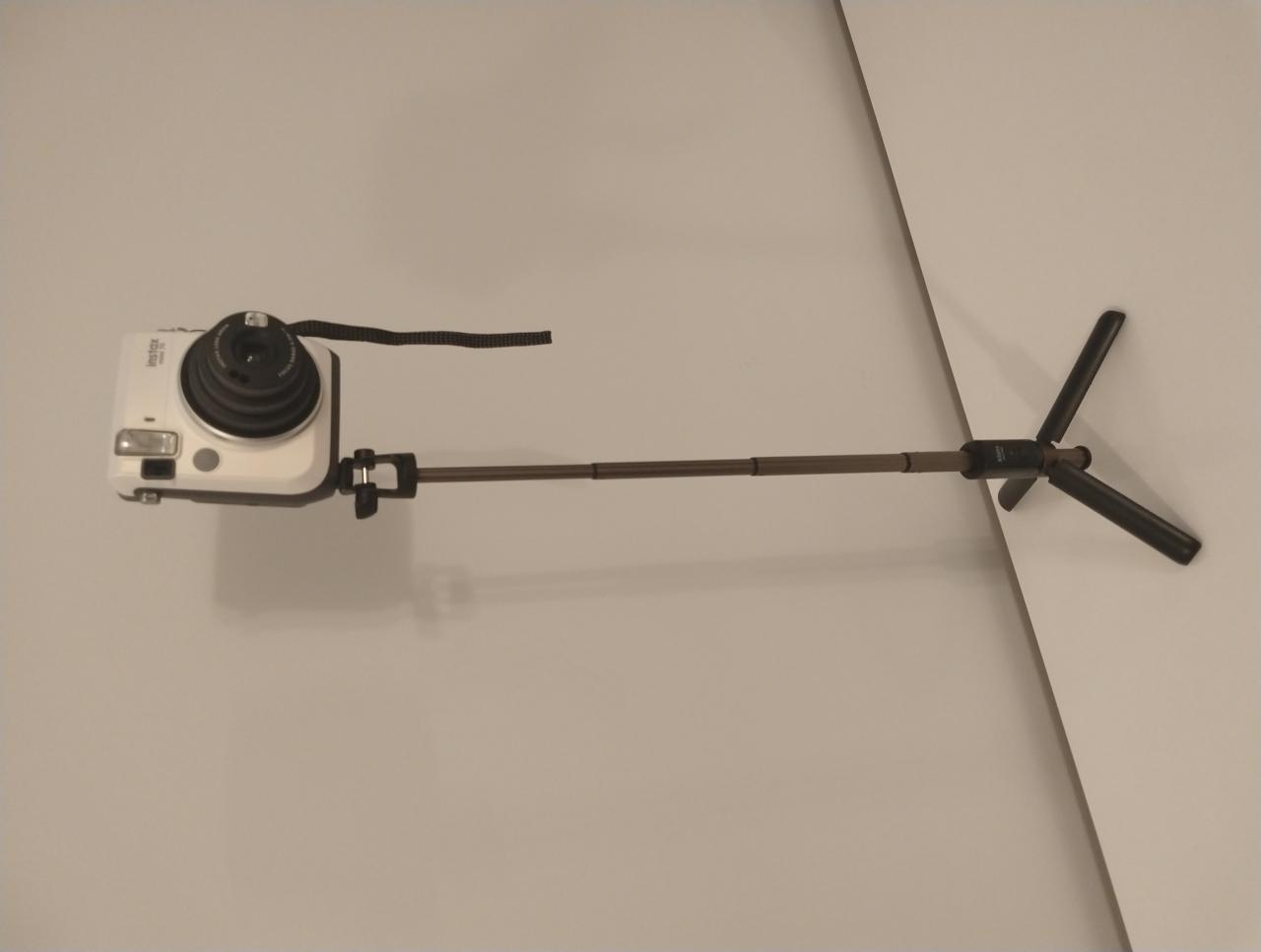 Biały aparat Instax przymocowany do brązowego teleskopowego statywu na tle jasnej ściany.