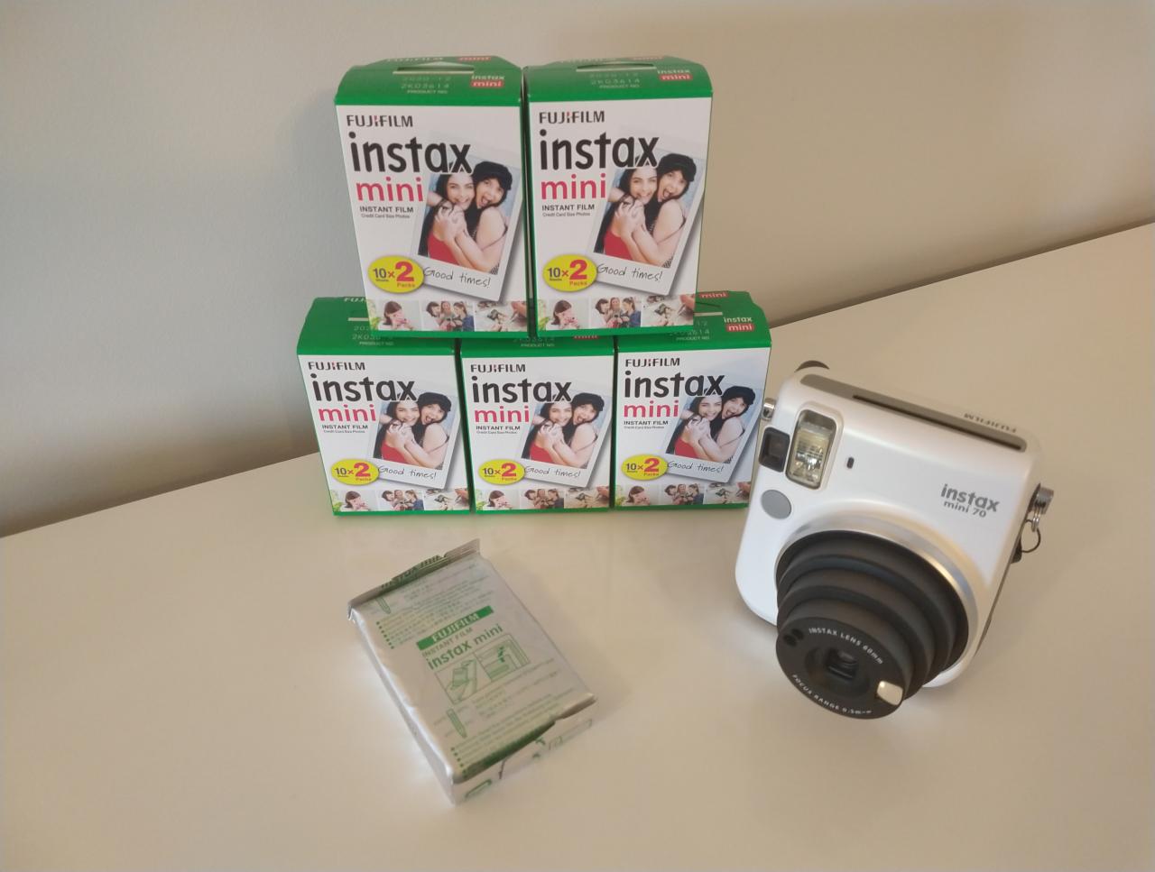 Biały aparat Fujifilm Instax Mini 70 obok opakowań wkładów Instax Mini na jasnym stole. Widoczny obiektyw aparatu i detale opakowań wkładów.