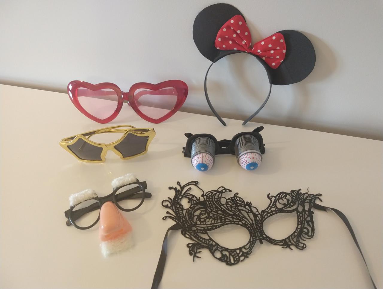 Różnorodny zestaw rekwizytów imprezowych na białym tle: okulary w kształcie serc, opaska z uszami Myszki Miki, okulary z gałkami ocznymi, okulary z nosem i wąsami, czarna maska karnawałowa.