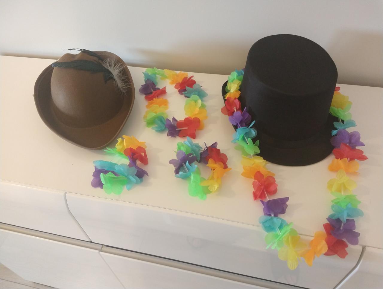 Dwa kapelusze, brązowy z piórkiem i czarny cylinder, oraz trzy hawajskie girlandy z kolorowych kwiatów leżące na białej komodzie.