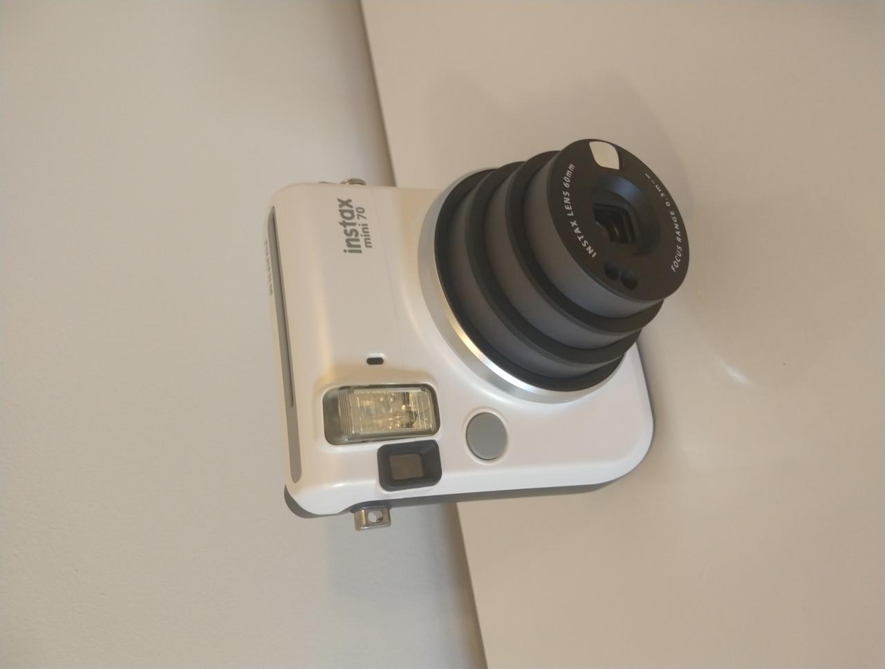 Biały aparat Instax mini 70 z obiektywem 60mm, leżący na jasnym tle.