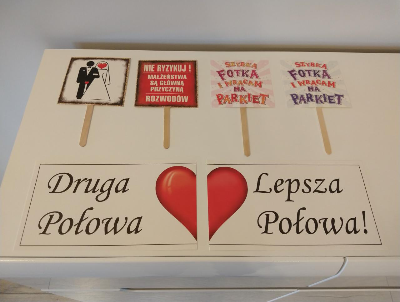 Dekoracje weselne z humorystycznymi napisami i grafikami, w tym 'Druga Połowa', 'Lepsza Połowa!', 'Szybka fotka i wracam na parkiet', oraz ostrzeżenie przed małżeństwem.