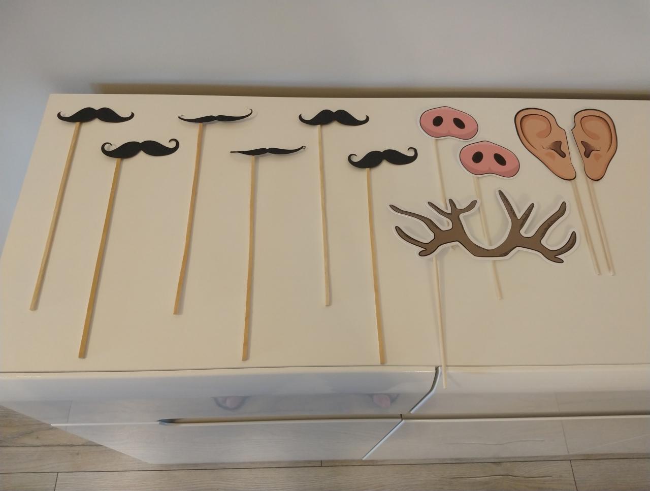 Różne rekwizyty fotograficzne na patyku, w tym wąsy, nosy, uszy i poroże, ułożone na białej komodzie.