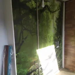 Przesuwne drzwi szafy z fototapetą przedstawiającą bujny, zielony las z wodospadem, oświetlone wiszącą żarówką i padającym światłem.