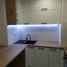 Nowoczesna kuchnia z białymi szafkami, drewnianym blatem i czarnym zlewozmywakiem, podświetlona listwą LED, z dekoracyjną mozaiką na ścianie i wzorzystą podłogą.