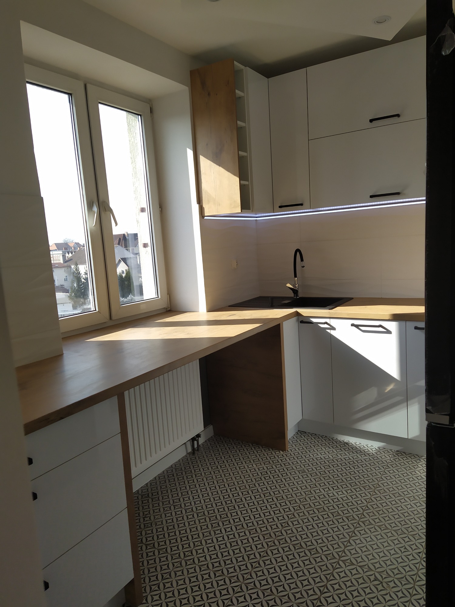 Jasna kuchnia z drewnianym blatem roboczym przy oknie, czarną armaturą i wzorzystą podłogą.