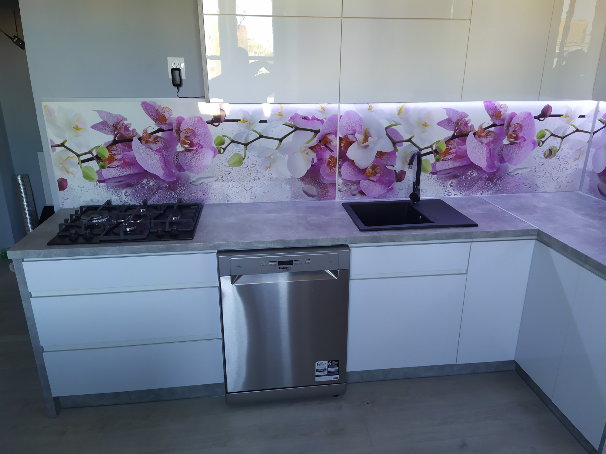 Nowoczesna kuchnia z białymi, lakierowanymi frontami, szarym blatem i panelem szklanym z motywem białych i fioletowych orchidei, z wbudowaną płytą gazową i zmywarką.