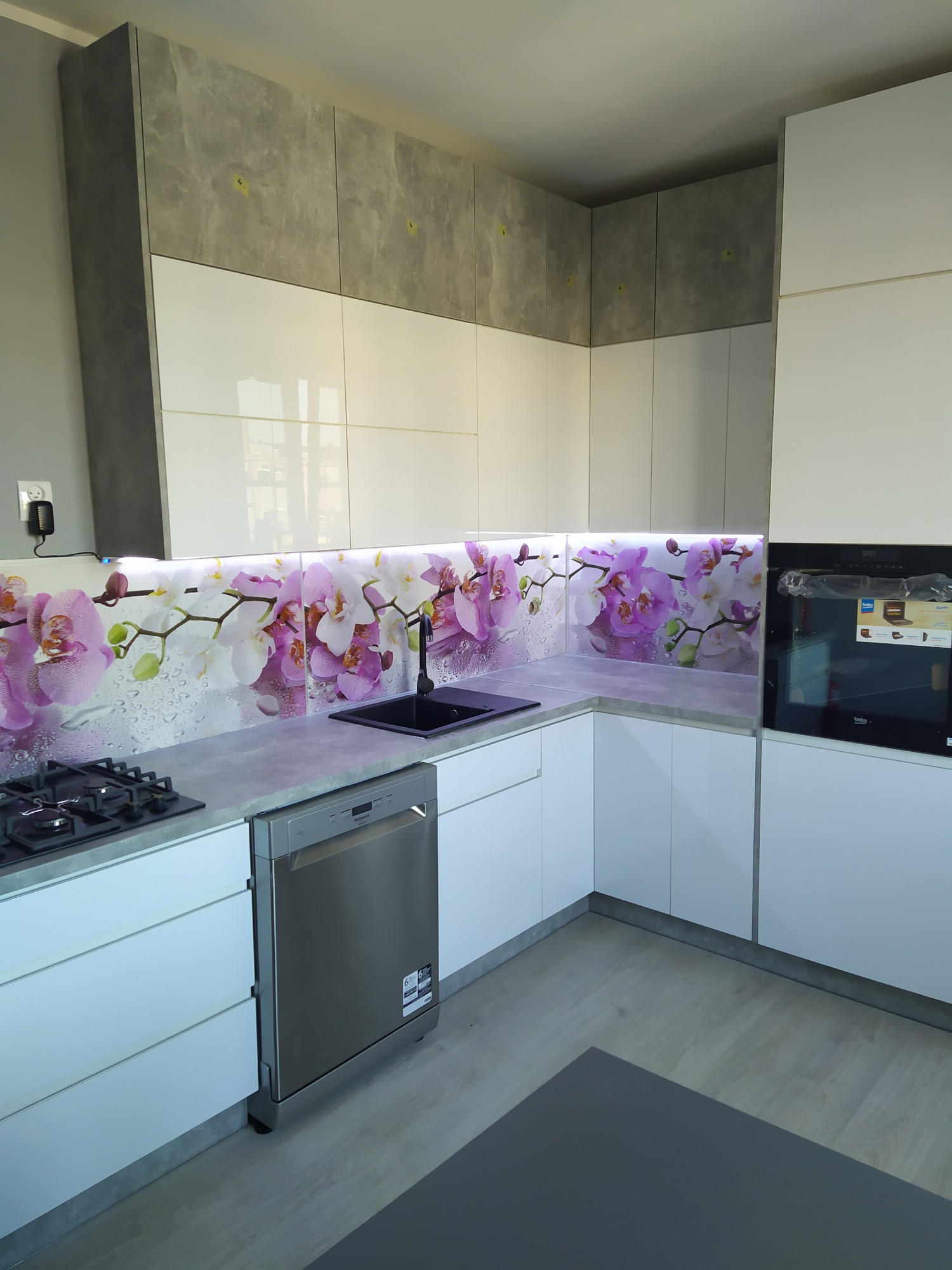 Nowoczesna kuchnia z białymi, lakierowanymi frontami szafek, betonowym blatem i panelem nad blatem z motywem białych i fioletowych orchidei, oświetlonym taśmą LED.