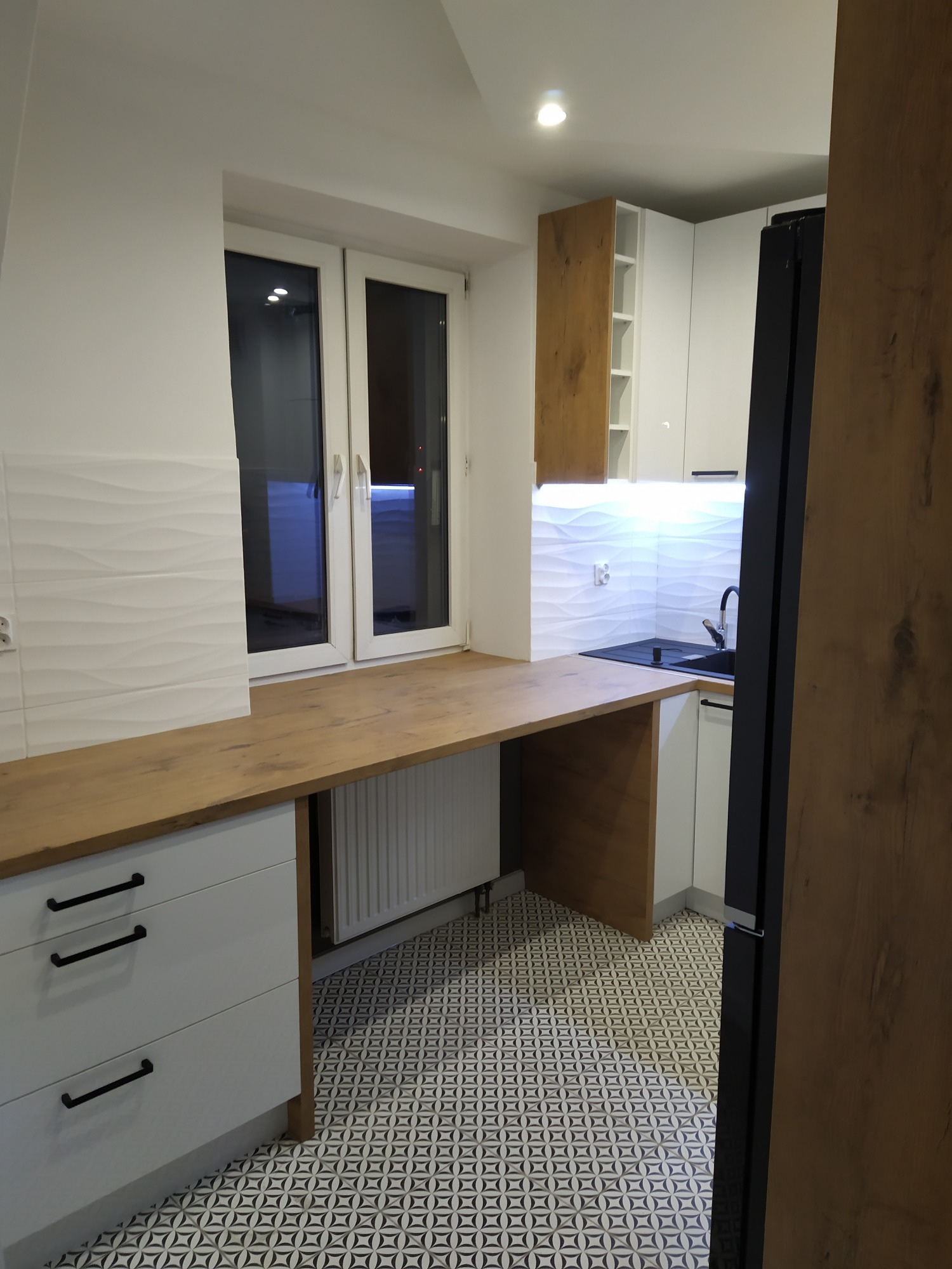 Nowoczesna kuchnia z drewnianym blatem roboczym pod oknem, białymi szafkami z czarnymi uchwytami i wzorzystą podłogą.