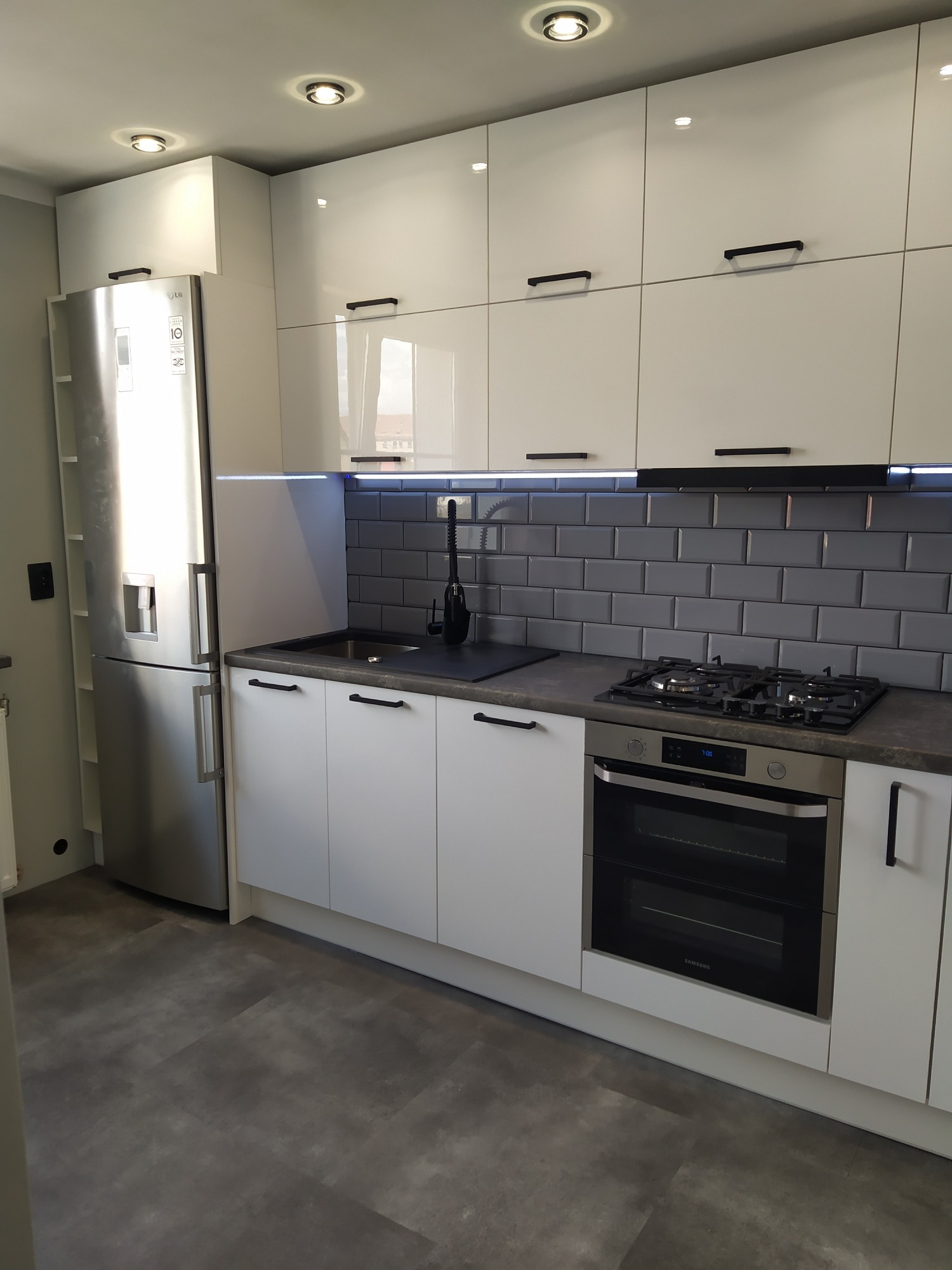 Nowoczesna kuchnia z białymi, lakierowanymi szafkami, grafitowym blatem i szarą cegłą na ścianie, z czarnymi, minimalistycznymi uchwytami i srebrną lodówką LG.