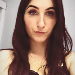 Portret kobiety o ciemnych włosach i jasnych oczach, z delikatnym makijażem i złotym naszyjnikiem w kształcie serca.