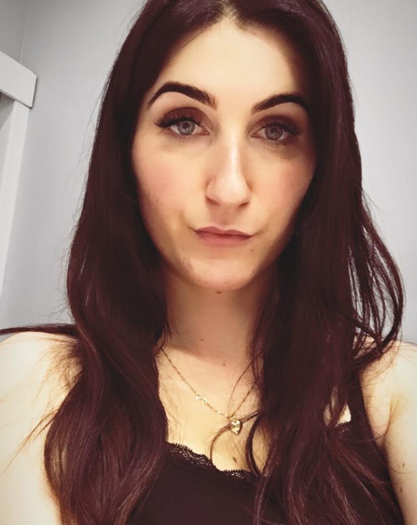 Portret kobiety o ciemnych włosach i jasnych oczach, z delikatnym makijażem i złotym naszyjnikiem w kształcie serca.