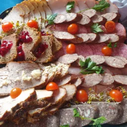 Dekoracyjnie ułożone na czarnym półmisku wędliny, pasztet z żurawiną, pieczone mięsa z sosem chrzanowym, kiełbasa, pomidorki cherry i kiełki.