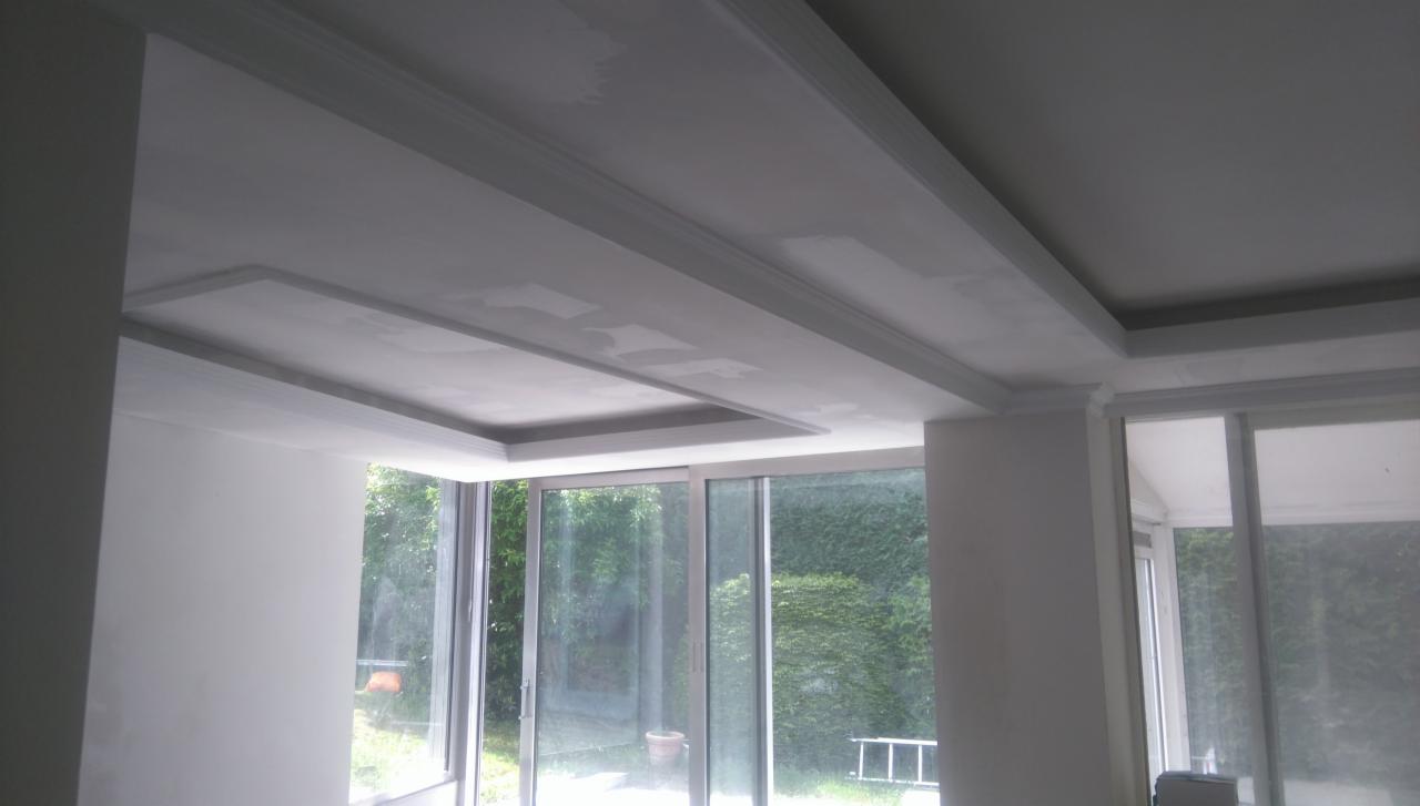 Niedokończony sufit z gładzią i dekoracyjnymi listwami w jasnym pomieszczeniu z dużymi oknami wychodzącymi na zieleń.