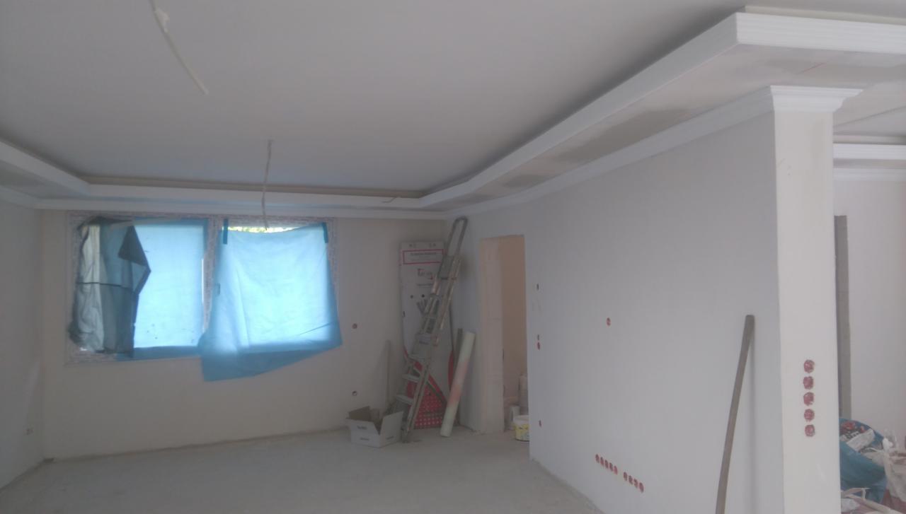 Wnętrze pokoju w trakcie prac wykończeniowych; okno zasłonięte folią ochronną, świeżo pomalowane ściany z listwami dekoracyjnymi pod sufitem, widoczna drabina i materiały budowlane.