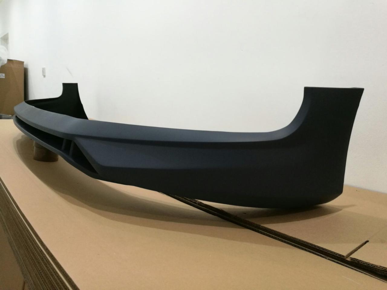 Formowany element z tworzywa sztucznego w kolorze grafitowym, prawdopodobnie obudowa lub część składowa większego urządzenia, leżący na tekturowej powierzchni, z widocznymi elementami wspornymi.