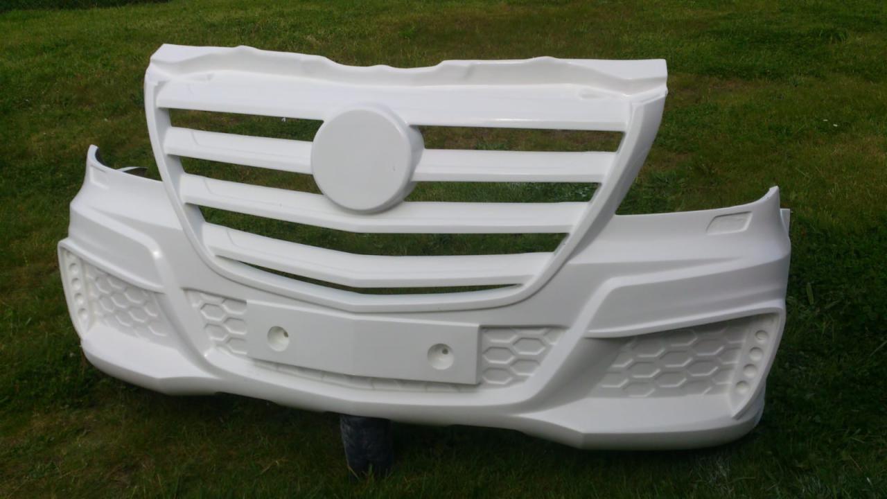 Nielakierowany, biały przedni zderzak samochodowy z atrapą grilla, leżący na trawie.