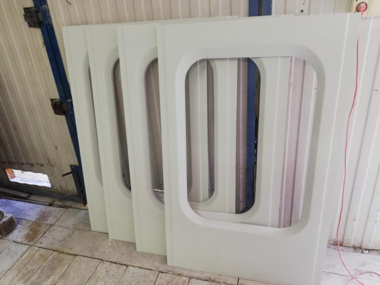 Białe, prostokątne ramy okienne z zaokrąglonymi rogami, ustawione rzędem w warsztacie, widoczne ślady obróbki.