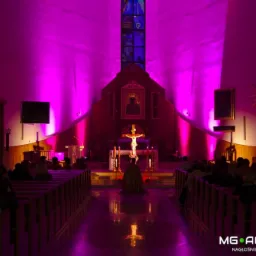 Kościół oświetlony fioletowym światłem, widoczny ołtarz i wierni siedzący w ławkach, na podłodze odbicie światła, w prawym dolnym rogu logo firmy MG Acoustics.