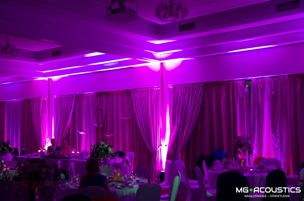 Sala bankietowa z dekoracjami kwiatowymi na stołach oświetlona fioletowym światłem, widoczne zasłony i żyrandole.