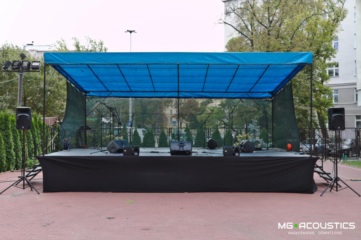 Scena plenerowa z niebieskim zadaszeniem, nagłośnieniem i mikrofonami, przygotowana do występu, widok z przodu na tle zieleni i budynku.