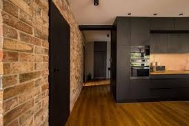 Loftowe wnętrze: ceglana ściana z czarnymi drzwiami, drewniana podłoga i ciemna zabudowa kuchenna z piekarnikiem w tle. Industrialny minimalizm.