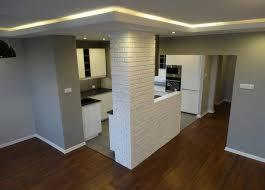 Nowoczesna kuchnia z drewnianą podłogą i szarą ścianą, oddzielona od reszty pomieszczenia niską ścianką z białej cegły, oświetlona listwami LED w suficie.