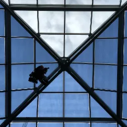 Pracownik w uprzęży, widoczny od dołu, czyści szklany dach budynku o konstrukcji kratownicowej, na tle błękitnego nieba z chmurami.