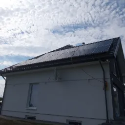 Dom jednorodzinny z zamontowaną instalacją fotowoltaiczną na dachu, widok z boku, z anteną satelitarną na ścianie i częściowo zachmurzonym niebem w tle.