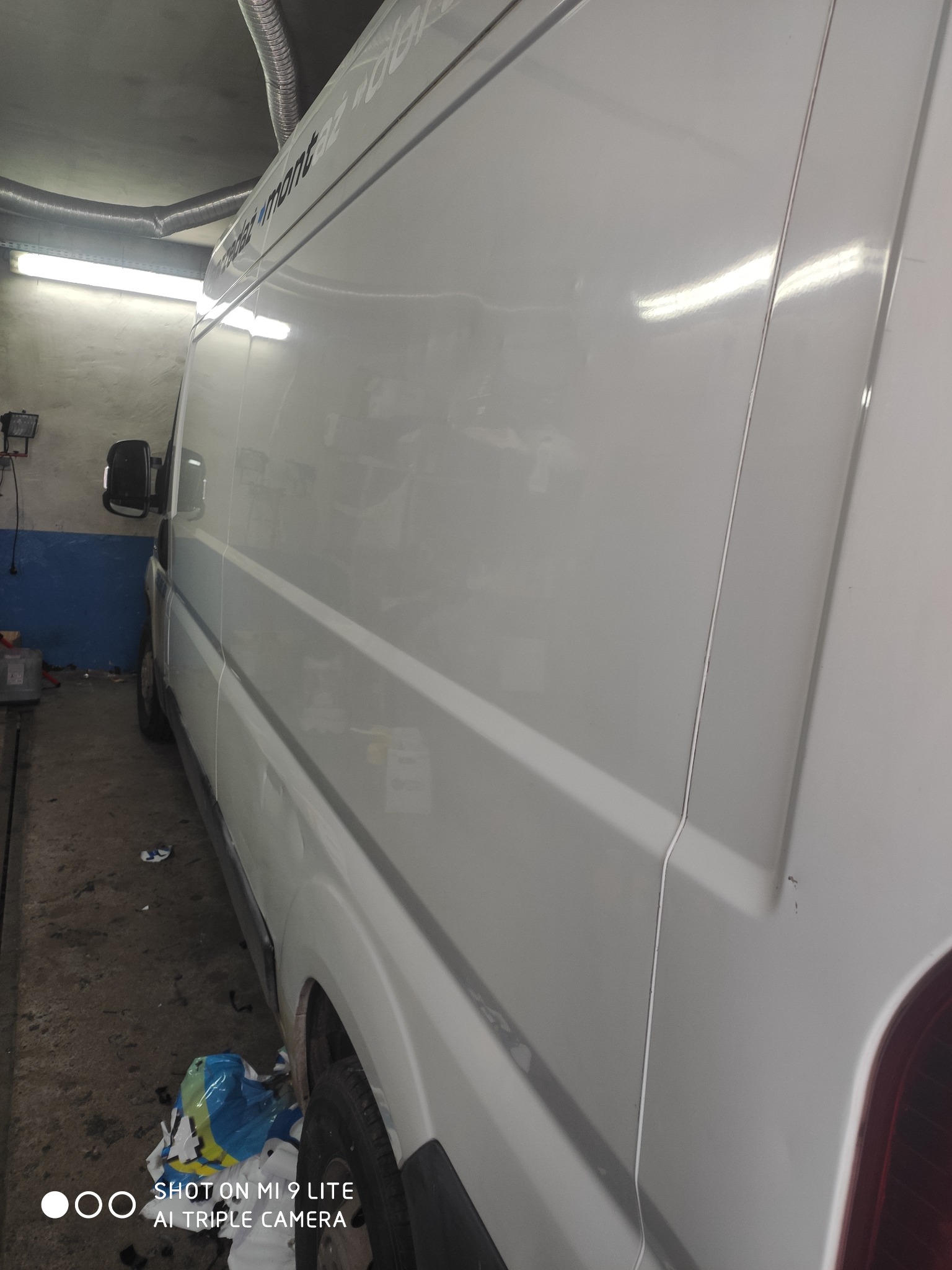 Biały bus marki Fiat Ducato w warsztacie, widok z boku, widoczny fragment napisu reklamowego na górze, oświetlenie warsztatowe odbija się w karoserii, na podłodze śmieci.